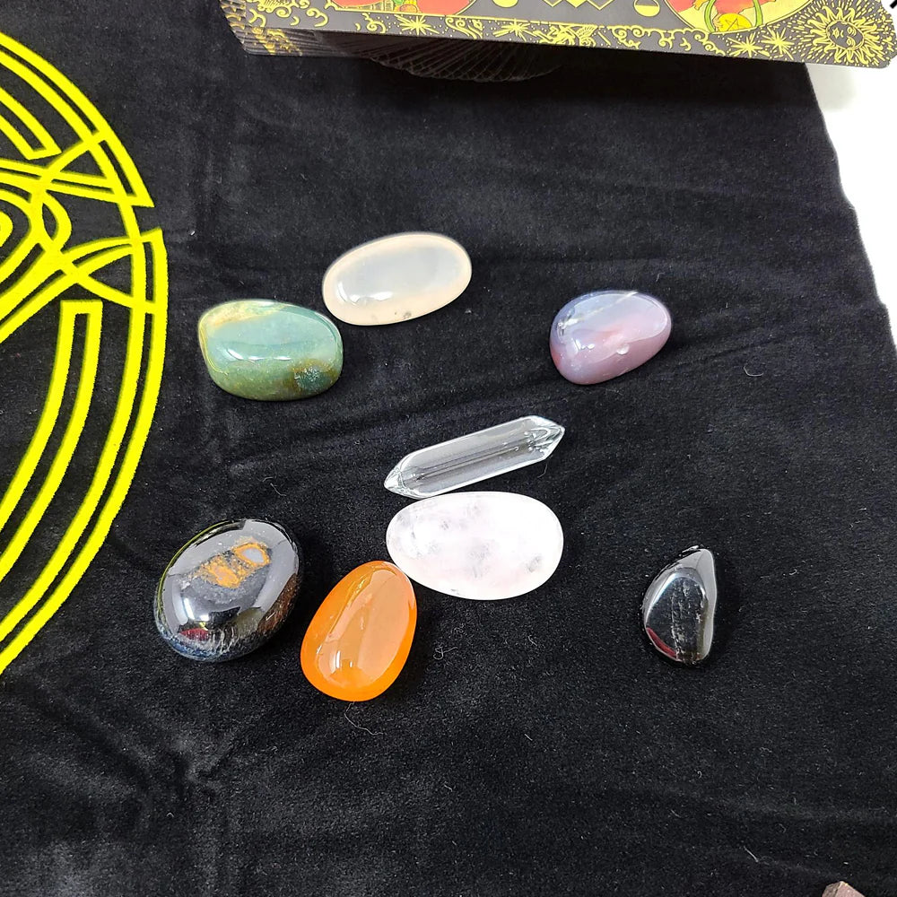 Variety of polished gemstones in different shapes and colors arranged on a black surface with a yellow circular pentagram design from the RWS tarot kit.