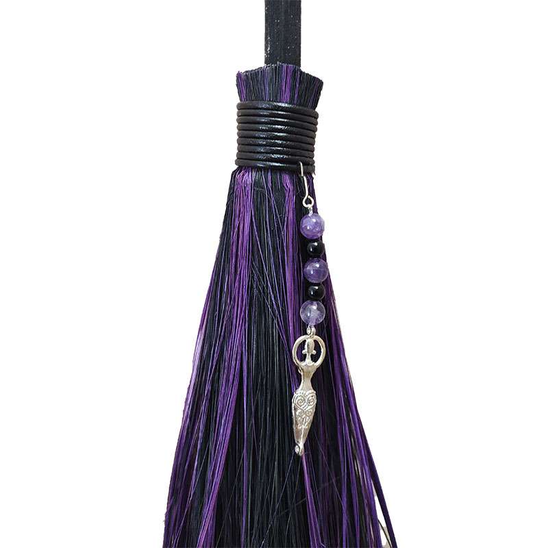 Close-up of the decorative top of a besom broom showing purple and black tassels, small beads, and a silver charm pendant.