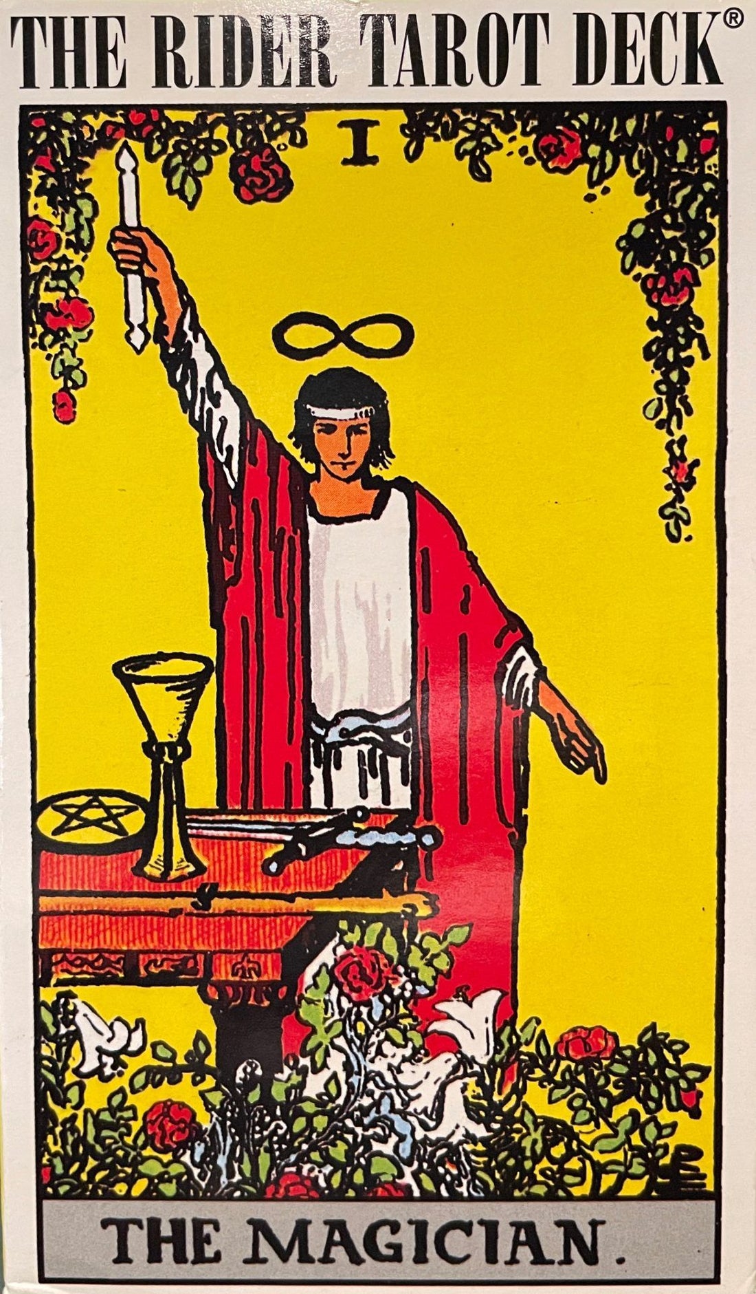 Rider-Waite Tarot Magician card showing a robed figure with one arm raised holding a wand above an altar bearing the four suit symbols.