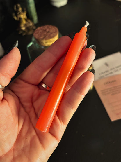 A hand with black nail polish holds a bright orange taper candle against a dark tabletop background with blurred decorative items.