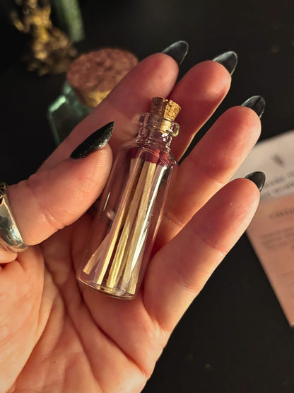 A hand with long black glittery nails holds a small clear glass bottle with a cork stopper containing gold-colored matchsticks, against a dark background.