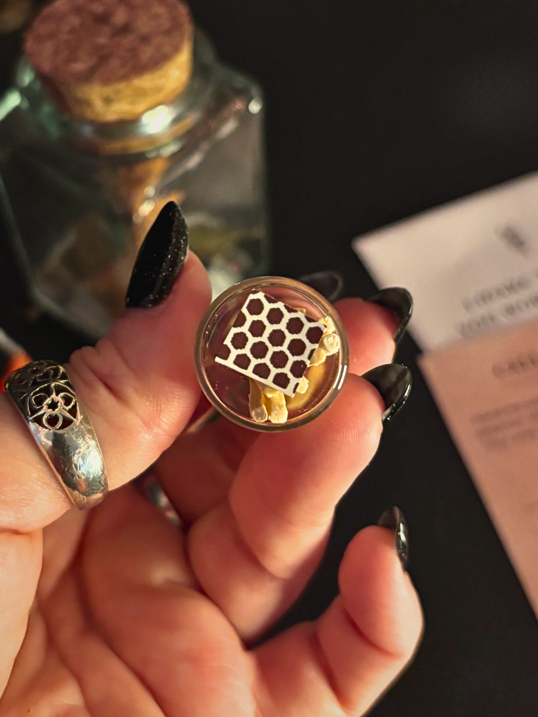 A hand with long black fingernails holds a small round glass vial containing a golden honeycomb structure and yellow wax pieces. A silver ring is worn on one finger, and a cork-topped glass bottle and a pink paper card are blurred in the background on a dark surface.