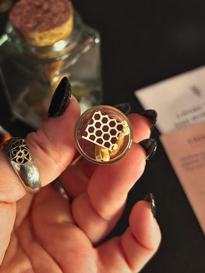 A hand with long black fingernails holds a small round glass vial containing a golden honeycomb structure and yellow wax pieces. A silver ring is worn on one finger, and a cork-topped glass bottle and a pink paper card are blurred in the background on a dark surface.