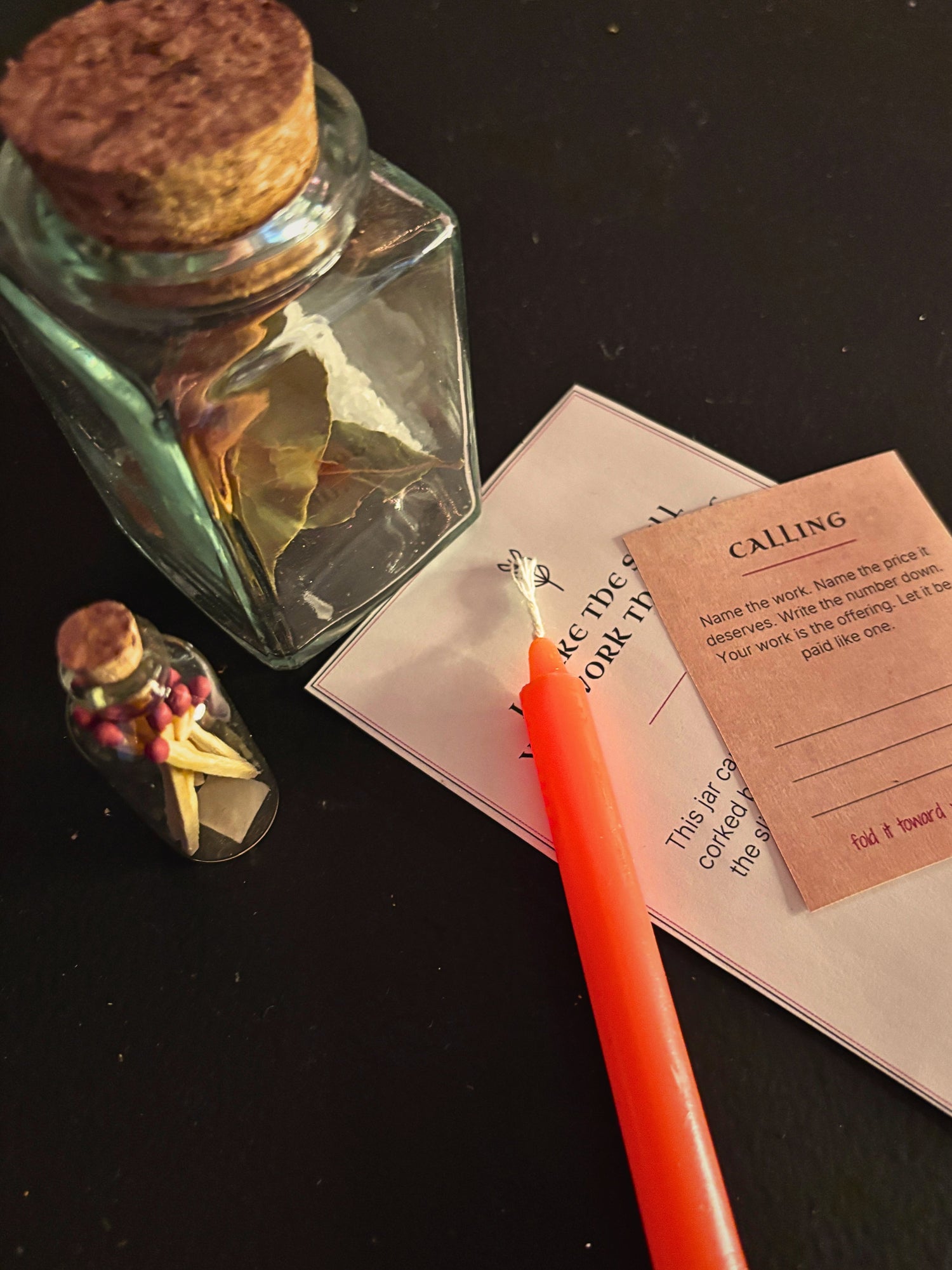 An orange taper candle with a silver star accent lies on top of a white card and a small kraft paper note on a dark surface. A large clear glass bottle with a cork stopper and a small glass vial are also visible in the scene.