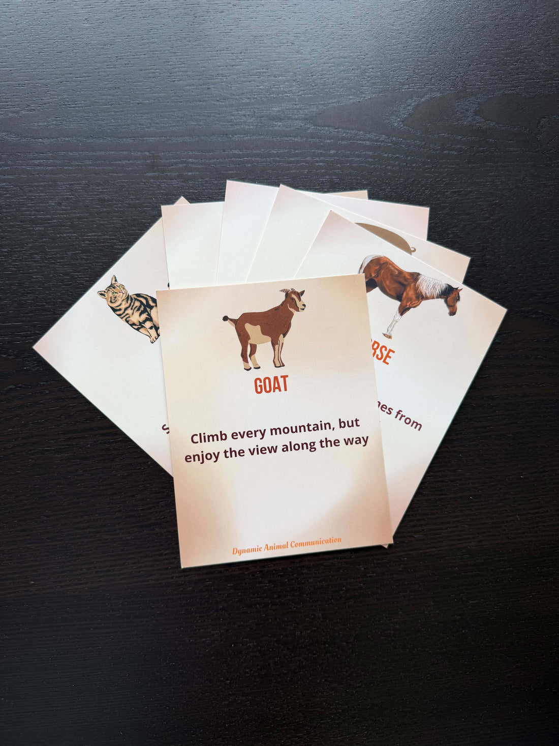 Multiple Farm Animal Wisdom Cards fanned out on a dark wood surface. The front Goat card reads &