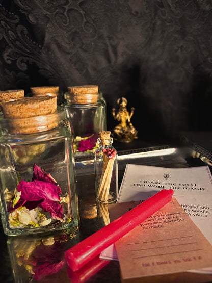 A styled tabletop arrangement featuring clear glass jars with cork stoppers, dried pink and white flowers, a small glass vial with a red wax seal, a red wax candle, a parchment note, and a printed card on a dark reflective surface.