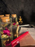 A styled tabletop arrangement featuring clear glass jars with cork stoppers, dried pink and white flowers, a small glass vial with a red wax seal, a red wax candle, a parchment note, and a printed card on a dark reflective surface.