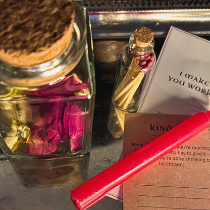 A close-up of a ritual or spell kit arranged on a metallic tray, including a glass bottle with a cork stopper and dried pink flowers, a small vial with a gold cap, printed cards or instructions, a brown paper slip, and a bright red wand.