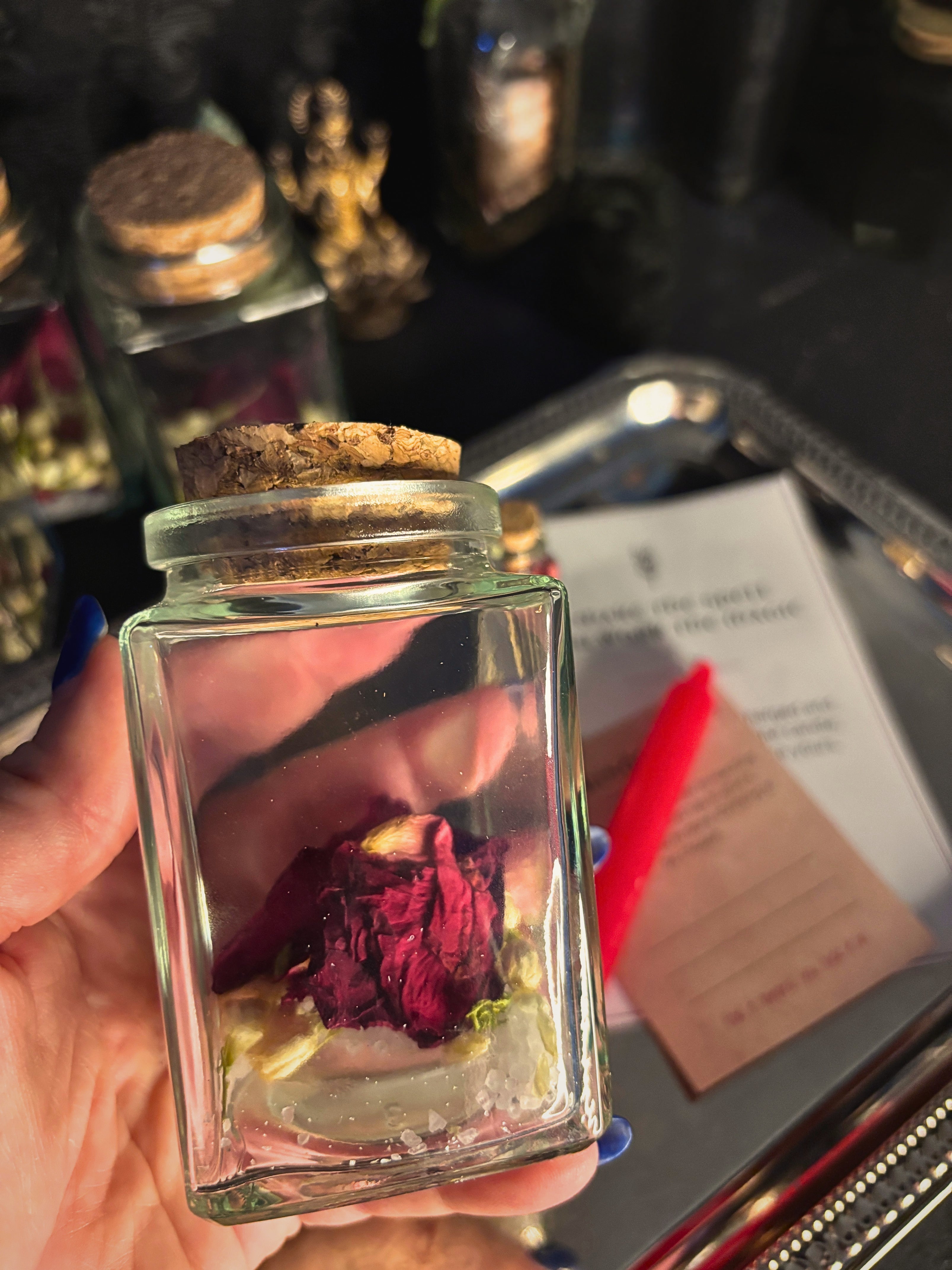 A hand holds a small clear glass jar with a cork stopper containing dried rose petals and white crystals, with other jars and cards in a dark setting in the background.