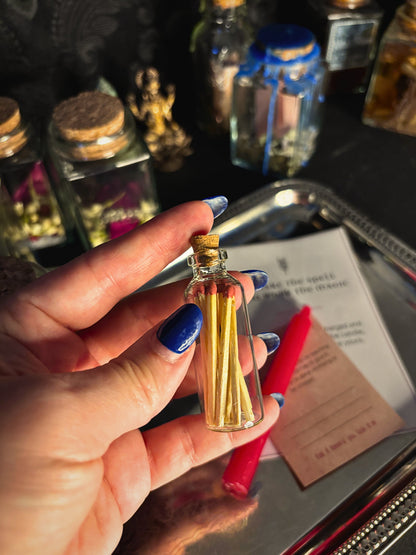 A hand with blue nail polish holds a small clear glass vial with a cork stopper containing yellow sticks, likely incense or香 sticks, over a tray with other decorative bottles and papers in a moody setting.