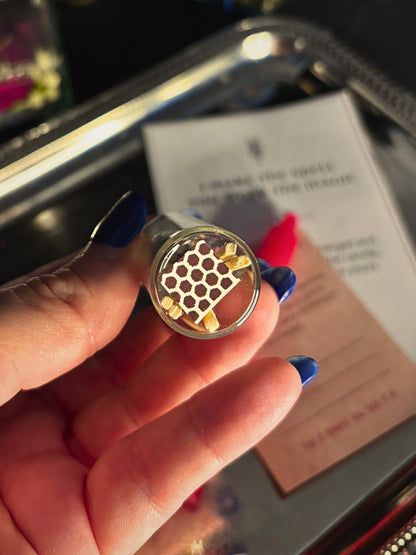 A hand with blue nail polish holds a small round metal object featuring a honeycomb pattern, with printed cards and a metallic tray blurred in the background.