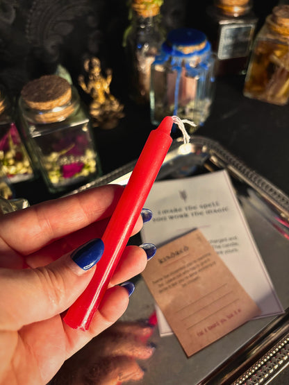 A hand with blue nail polish holds a bright red wax candle above a tray containing a card and other decorative jars in a dim, moody setting.
