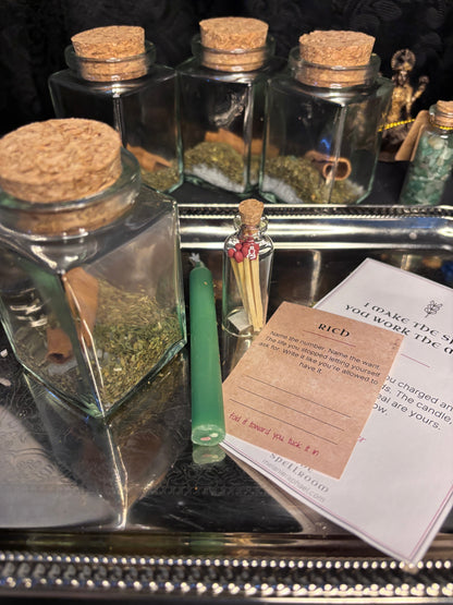 A styled tabletop arrangement of clear glass jars with cork stoppers containing dried herbs, a small vial with matches, a green glass tube, and printed cards on a reflective silver tray.