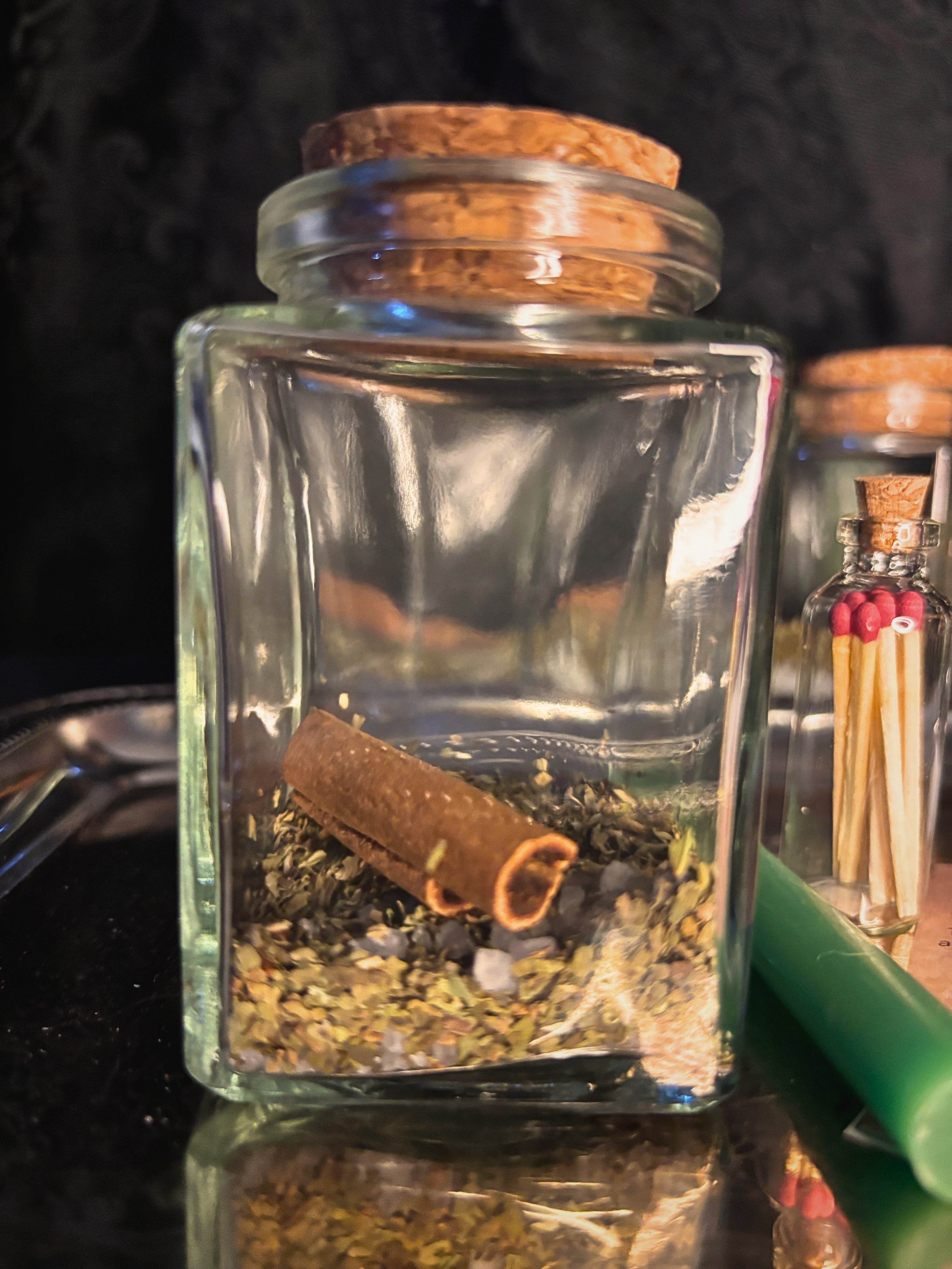 A clear glass jar with a cork stopper containing dried herbs and a cinnamon stick, photographed on a dark reflective surface with a blurred matchbox-style bottle in the background.