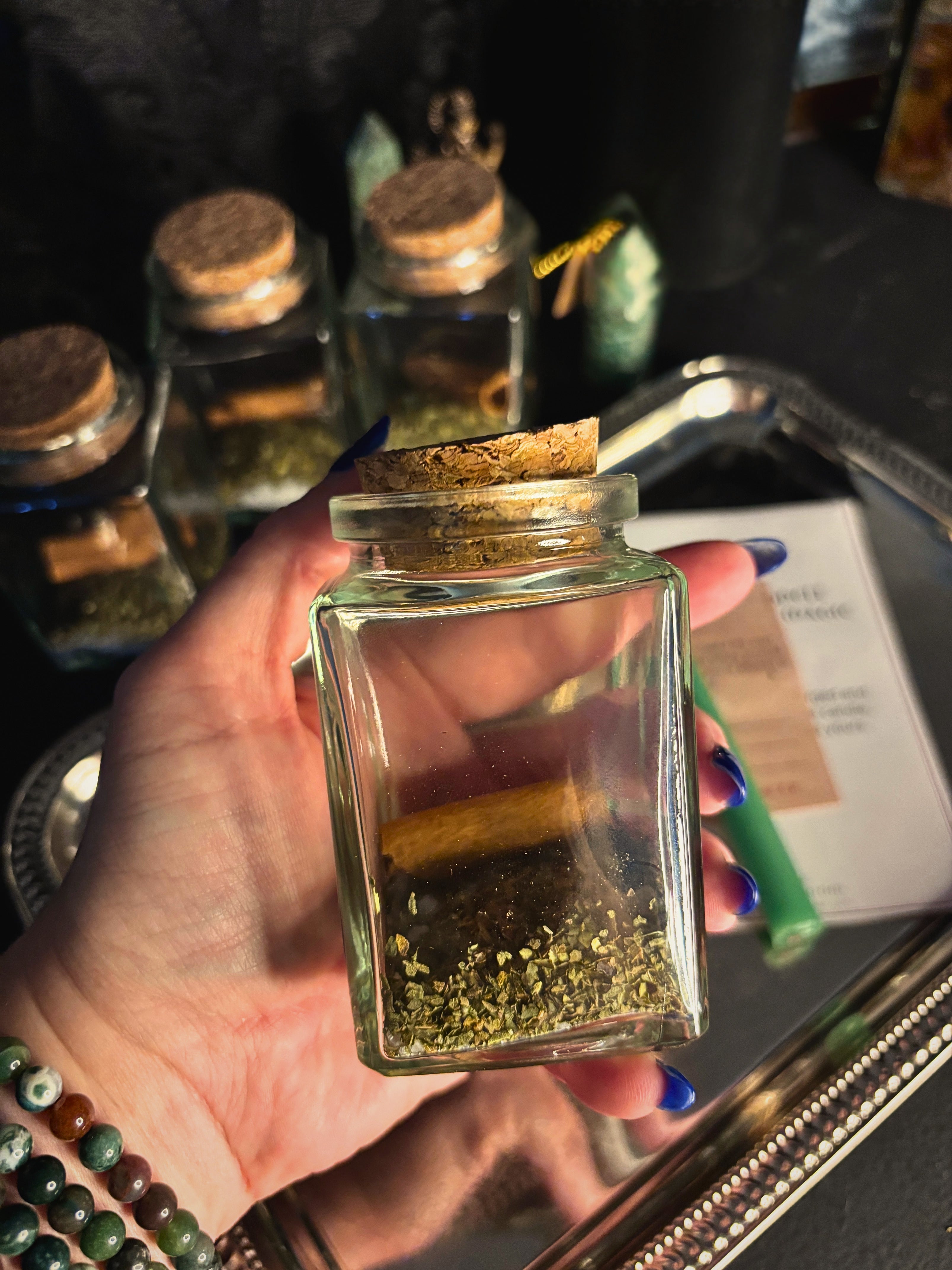 A hand holds a small clear glass jar with a cork stopper containing dried green herbs and a small gold piece, with other similar jars on a tray in the background.