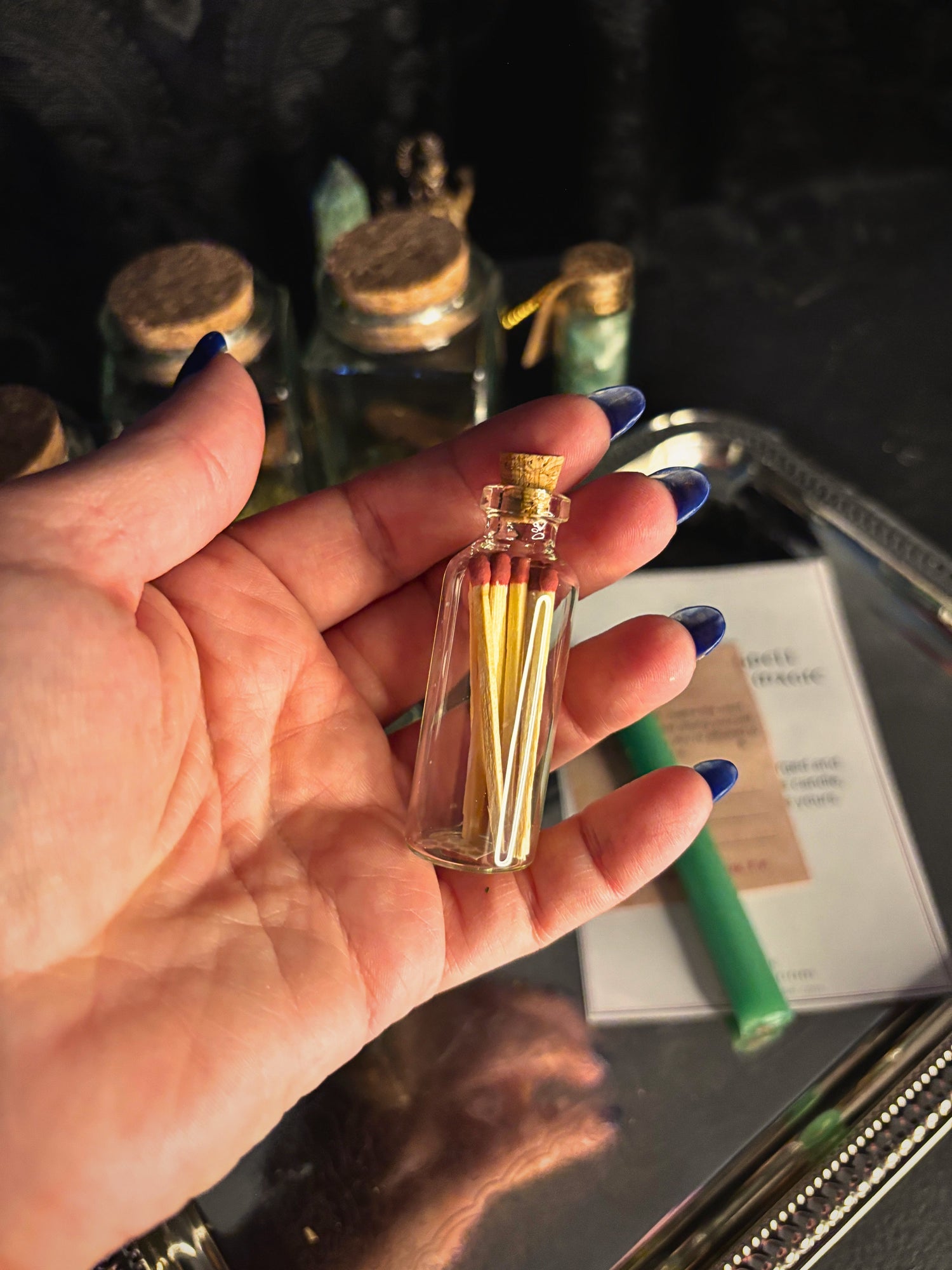 A hand with blue nail polish holds a miniature clear glass vial containing small golden-yellow matches with red tips, set against a dark, moody background with other glass bottles and a tray.