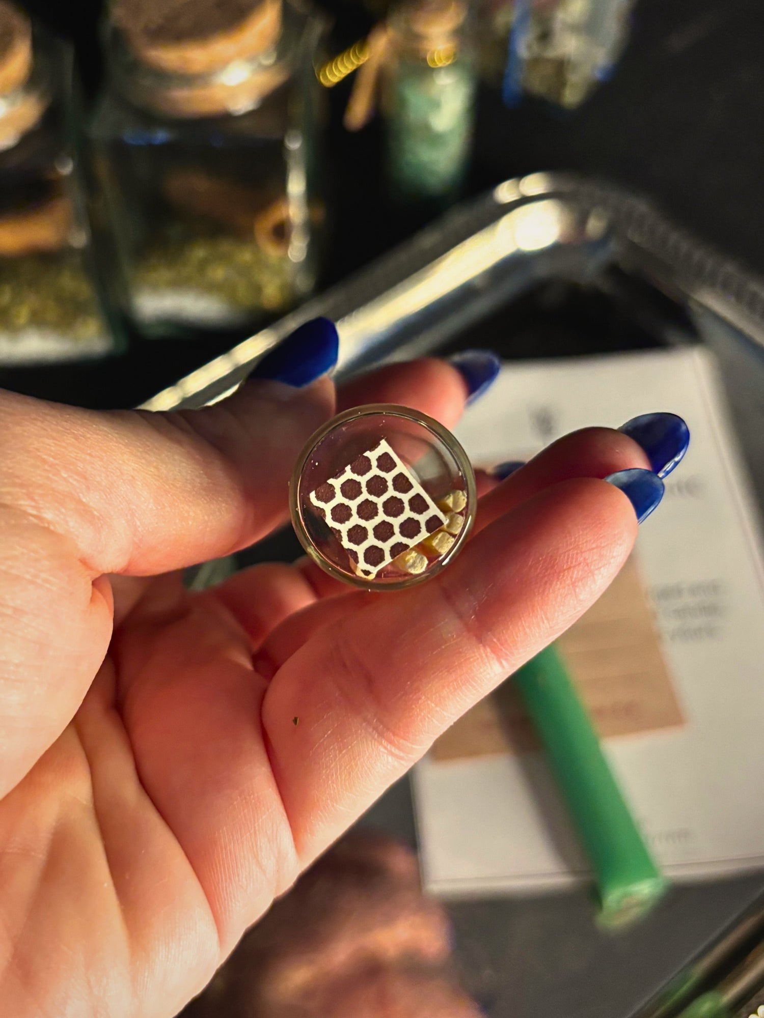 A hand with blue nail polish holds a small round glass jar containing a gold honeycomb structure and tiny gold beads. The background is a dark, softly blurred setting with glass bottles and a metallic tray.