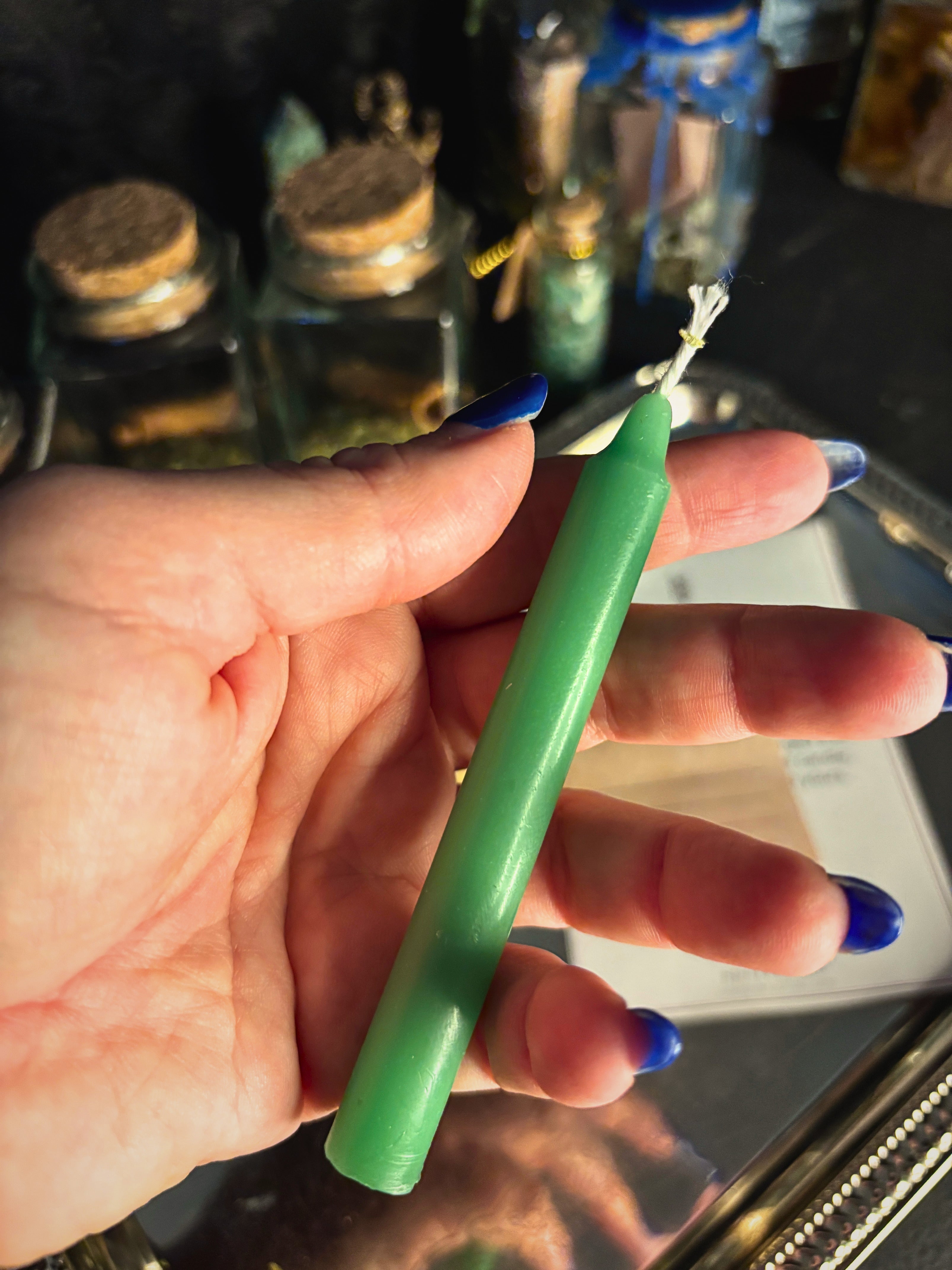 A hand with blue nail polish holds a solid green taper candle with a white wick, set against a dark, blurred background with glass jars.
