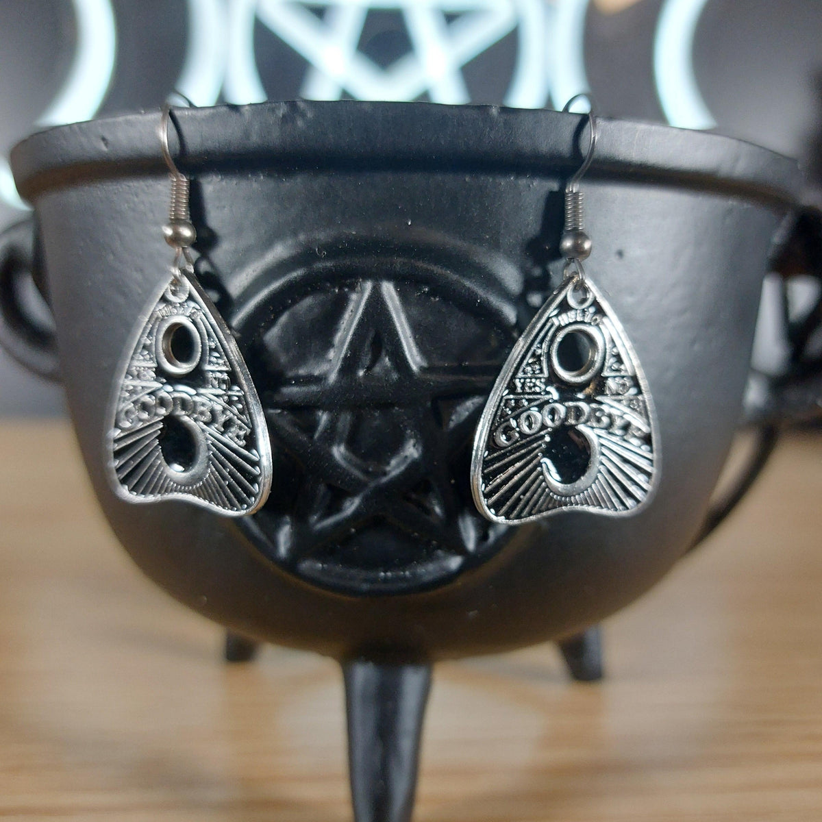 Silver tone Ouija planchette "Goodbye Yes No" dangle earrings on a black cauldron.