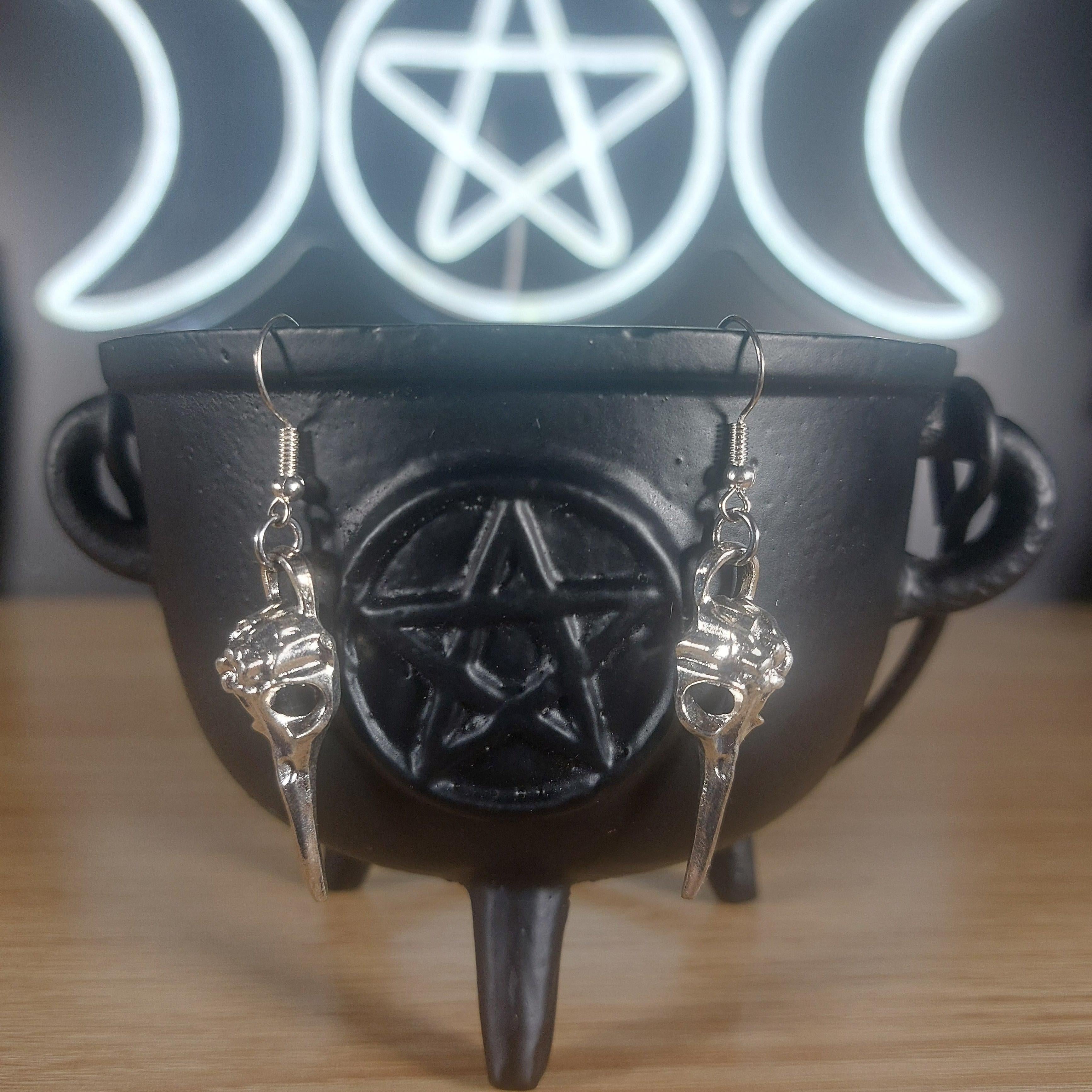 Silver-tone raven skull dangle earrings hanging from a black cauldron.