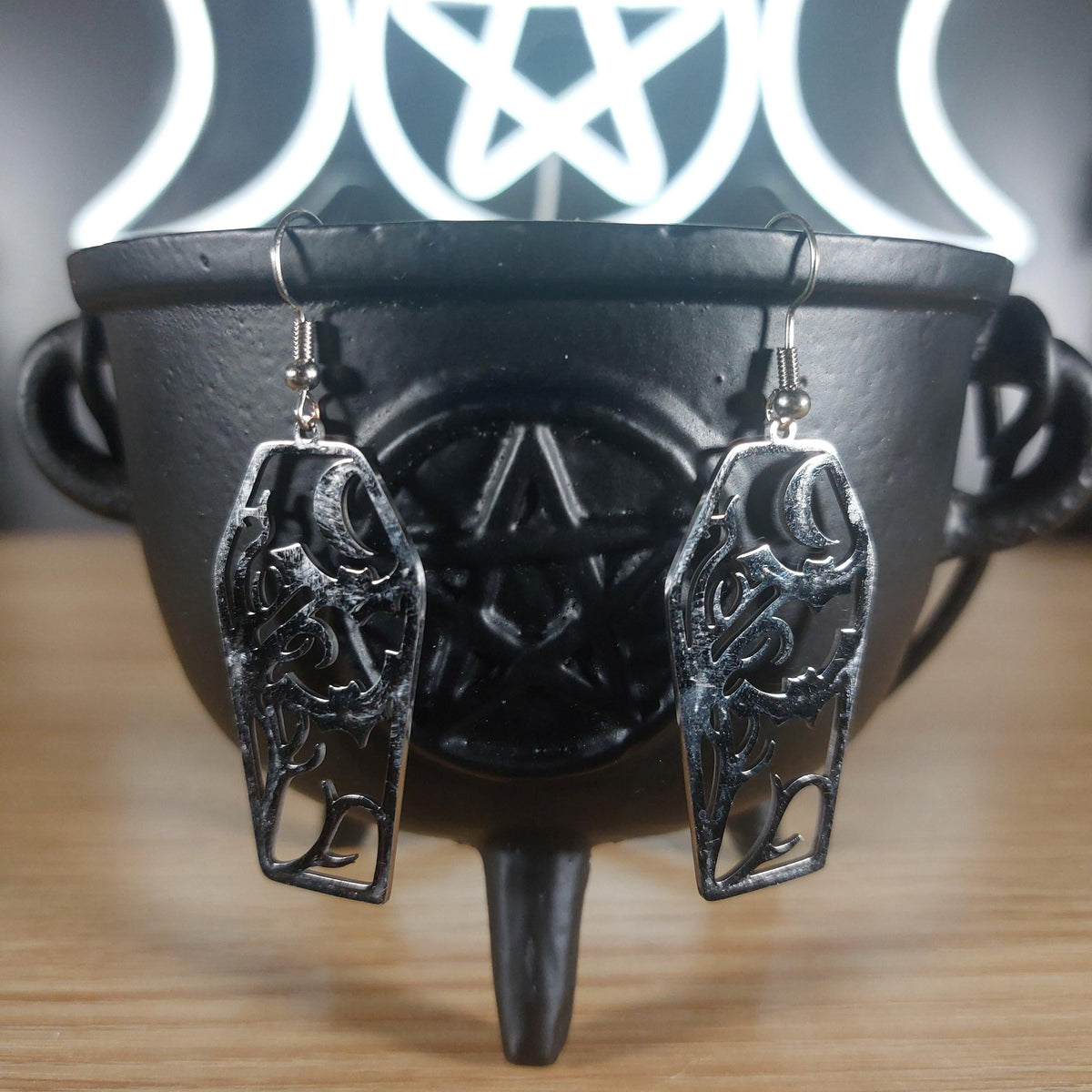 Silver tone coffin dangle earrings with cutout bats and moon design, hanging from a black cauldron.