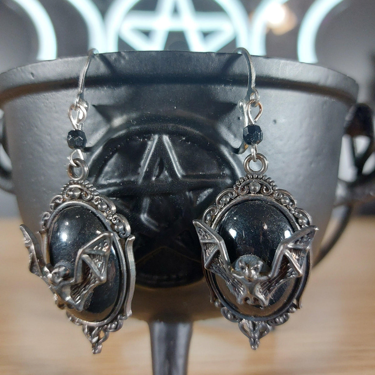 Antique silver bat dangle earrings with black oval cabochons, hanging from a black cauldron.
