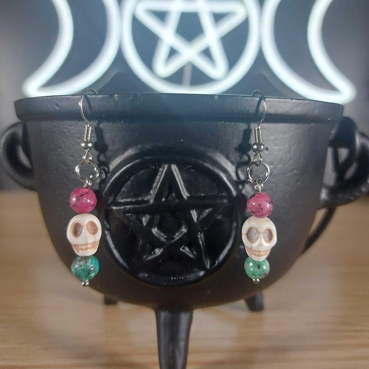 White howlite skull dangle earrings with pink and green glass beads on a black cauldron.