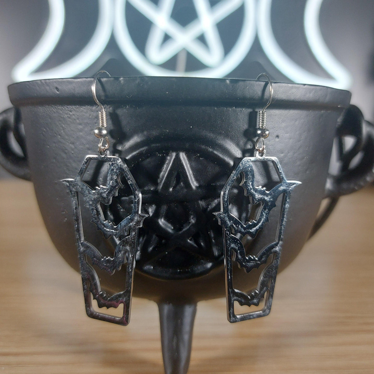 Silver tone coffin dangle earrings with cutout bats, resting on a black cauldron.