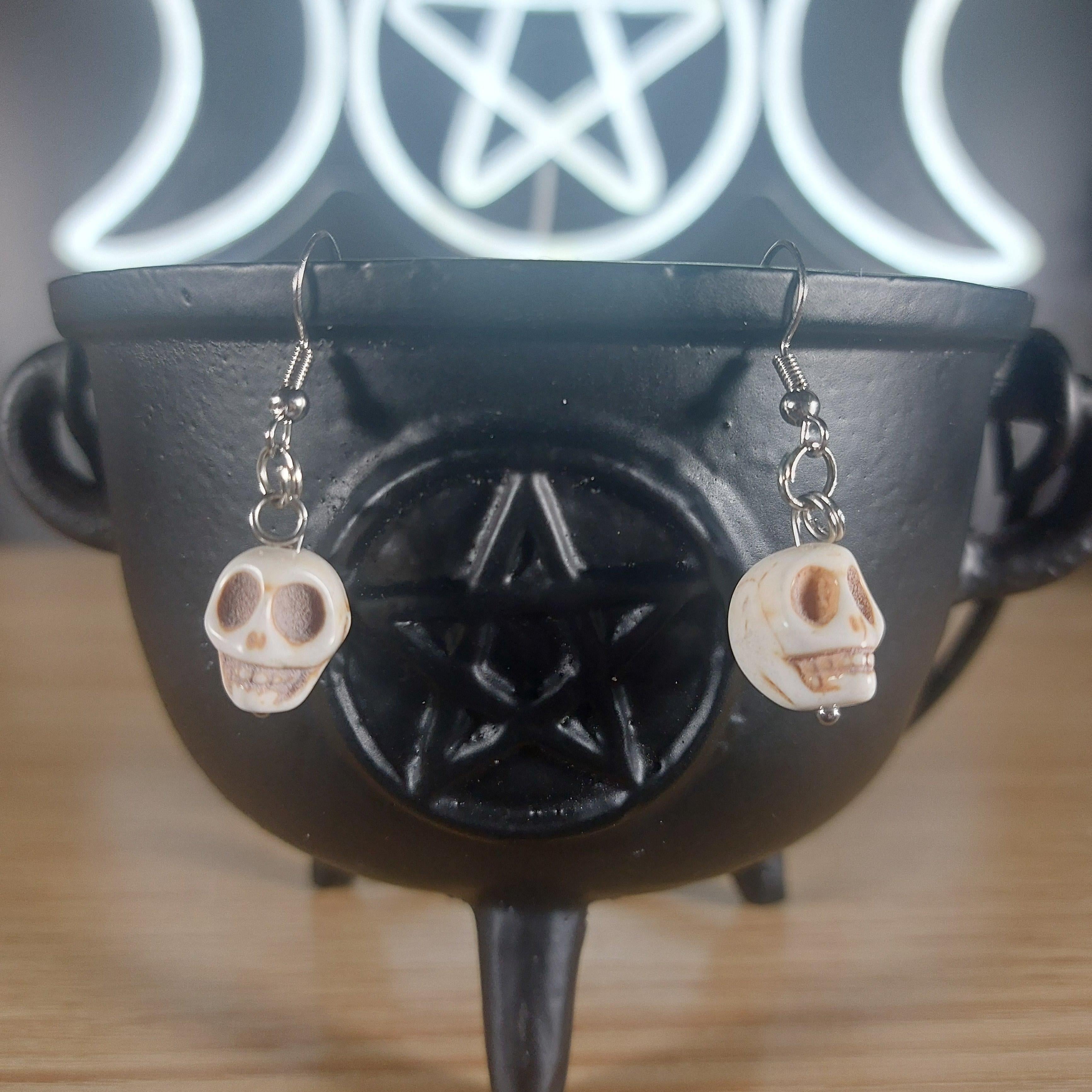 White howlite crystal dangle earrings with skull charms, hanging from a black cauldron.
