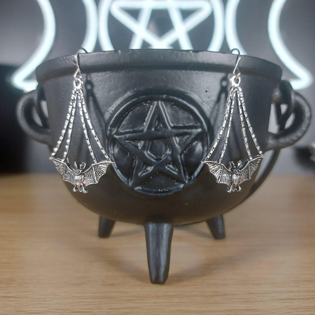 Antique silver dangle bat earrings hanging from a black cauldron with a pentagram design.