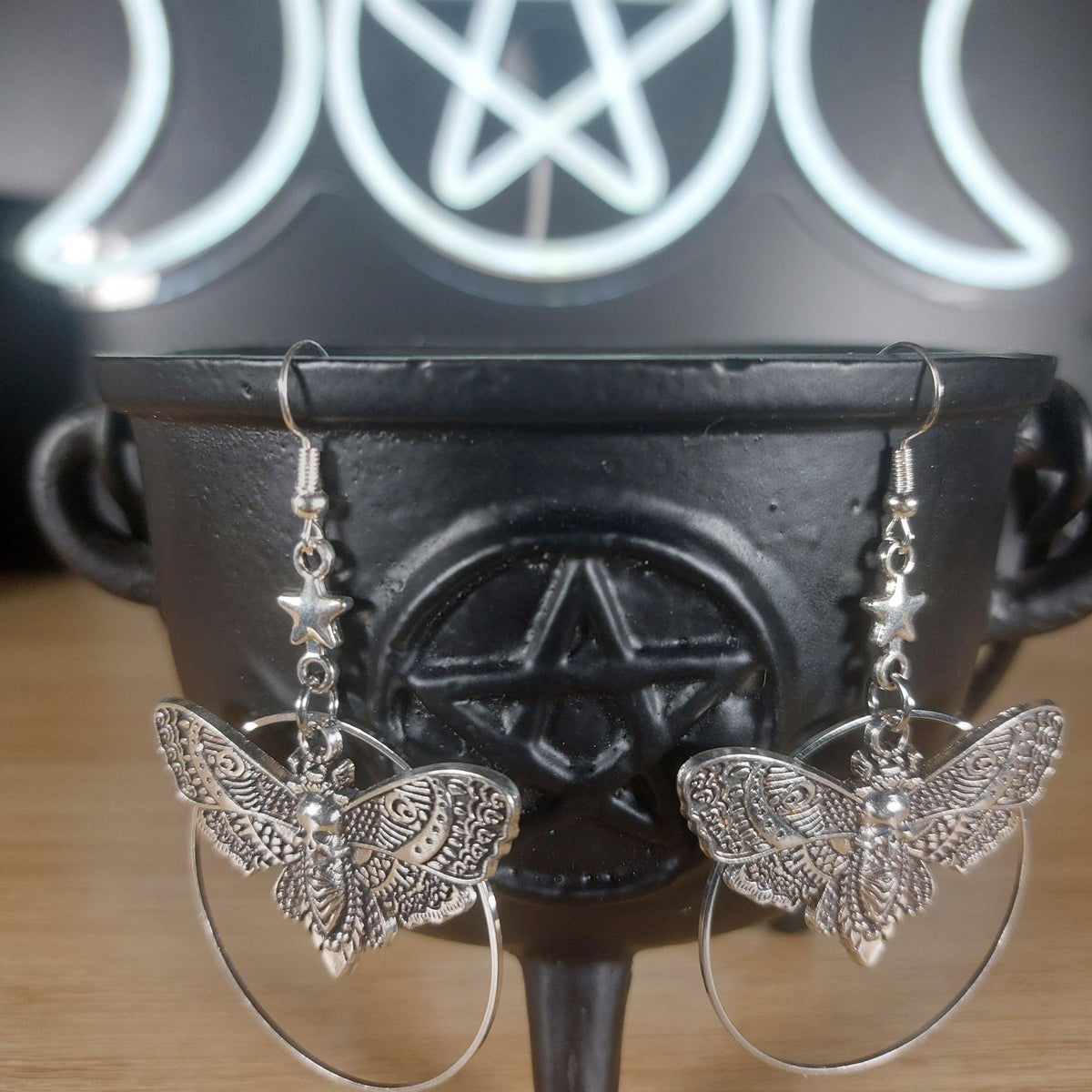Silver tone death moth dangle earrings with star accents and hoop details, resting on a black cauldron.