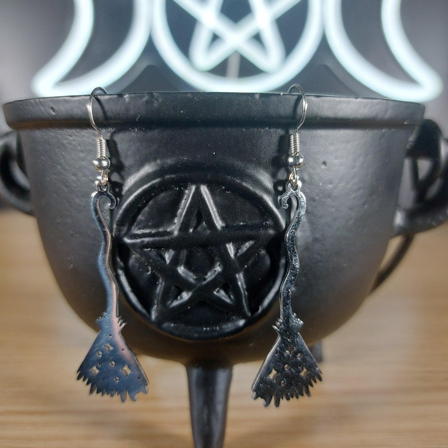 Silver tone broom dangle earrings hanging from a black cauldron with a pentagram.