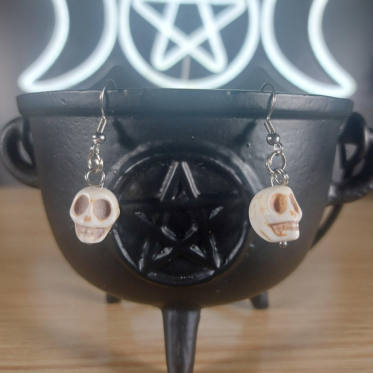 White howlite crystal dangle earrings with skull charms, hanging from a black cauldron.