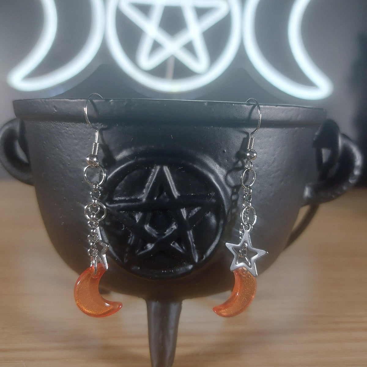 Orange glass moon and silver star dangle earrings hanging from a black cauldron.