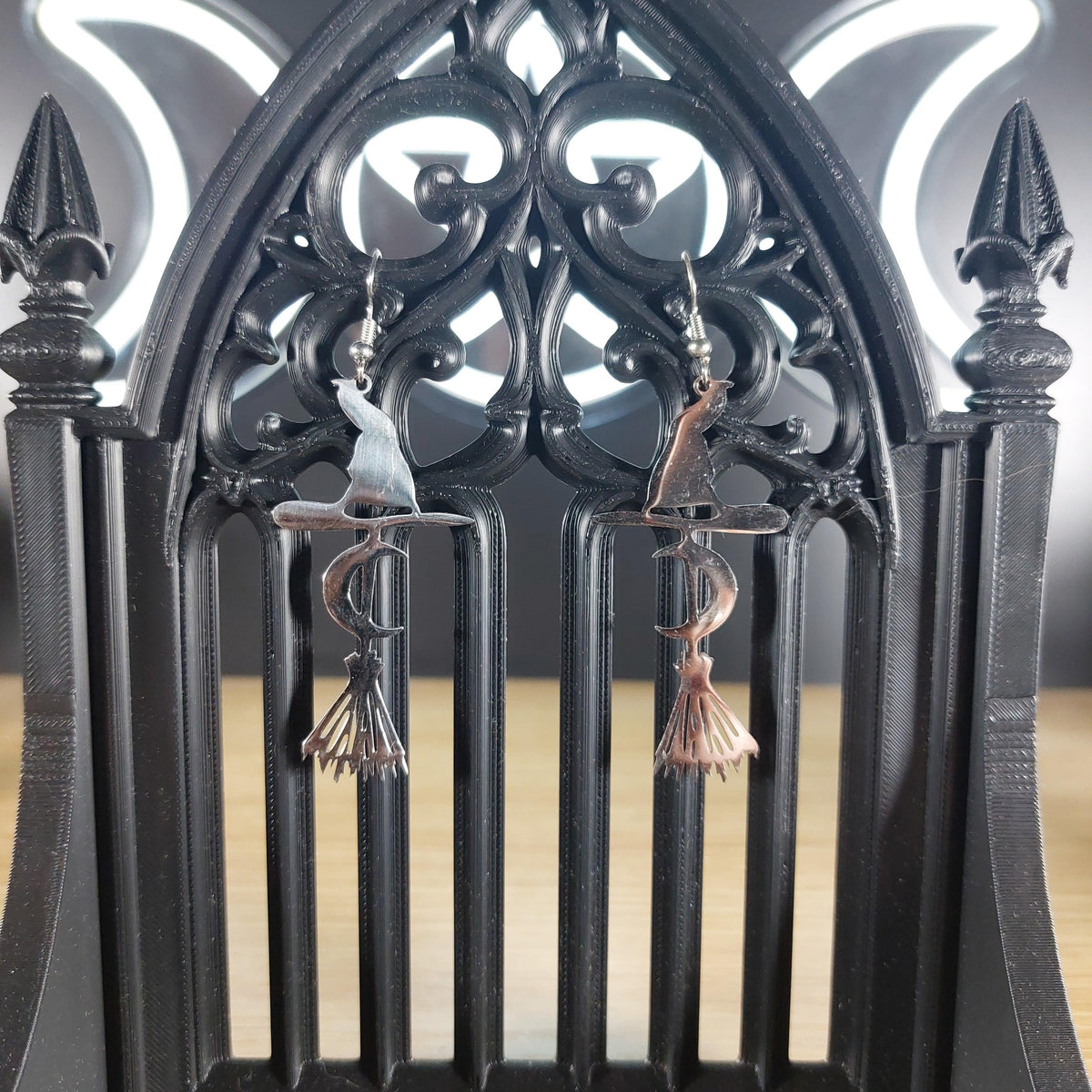 Silver tone broom, witch hat, and crescent moon dangle earrings on a black gothic arch display.