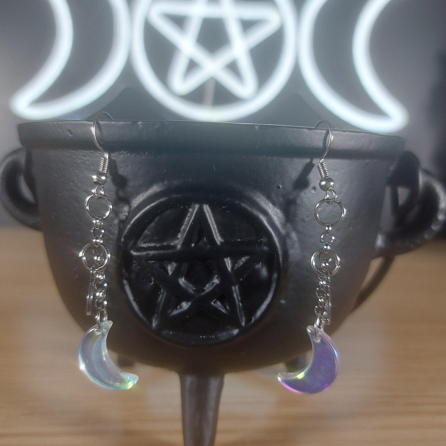 Iridescent clear glass moon dangle earrings hanging from a black cauldron with a pentagram.