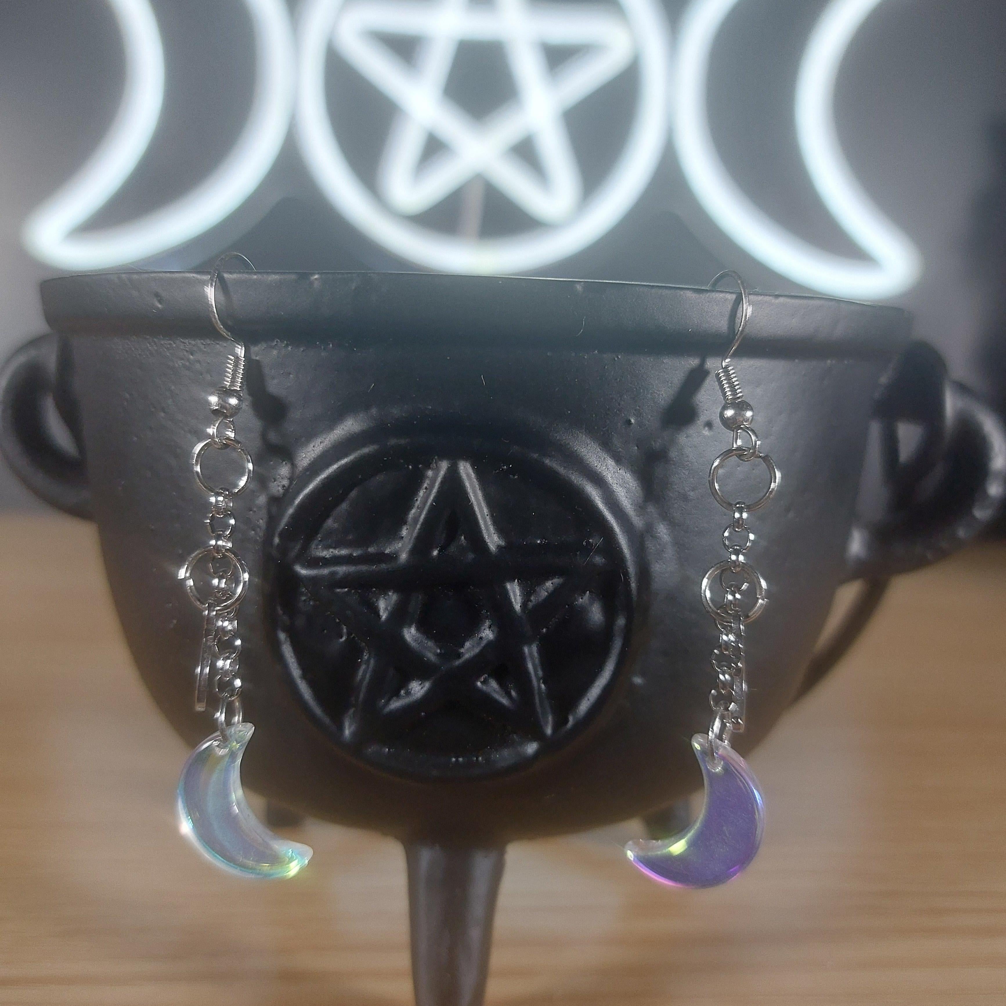 Iridescent clear glass moon dangle earrings hanging from a black cauldron with a pentagram.