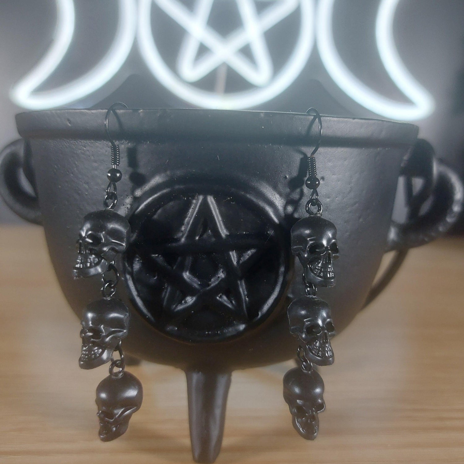Black plastic 3-tier skull dangle earrings on a black cauldron with a glowing pentagram and triple moon symbol.