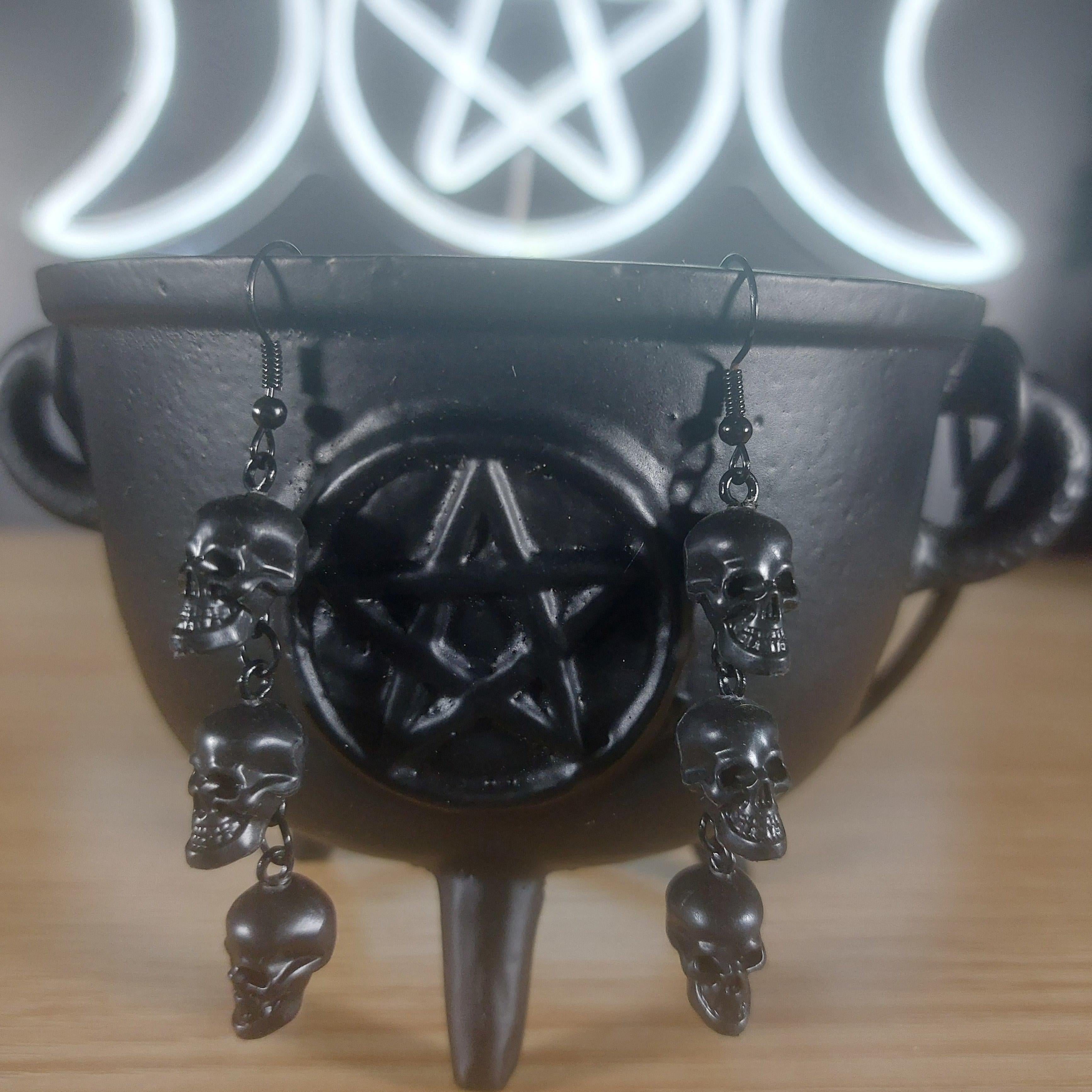 Black plastic 3-tier skull dangle earrings on a black cauldron with a glowing pentagram and triple moon symbol.