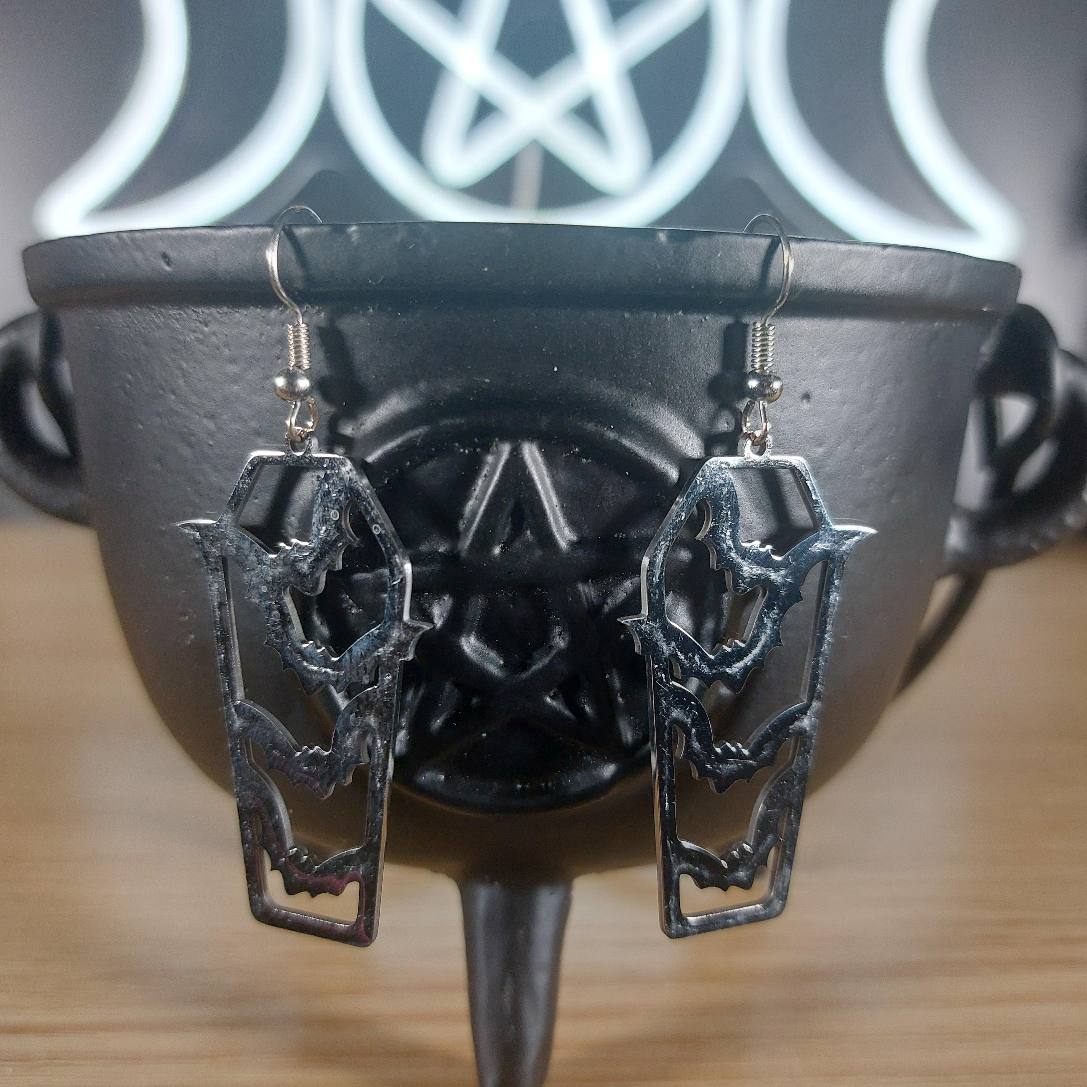 Silver tone coffin dangle earrings with cutout bats, resting on a black cauldron.