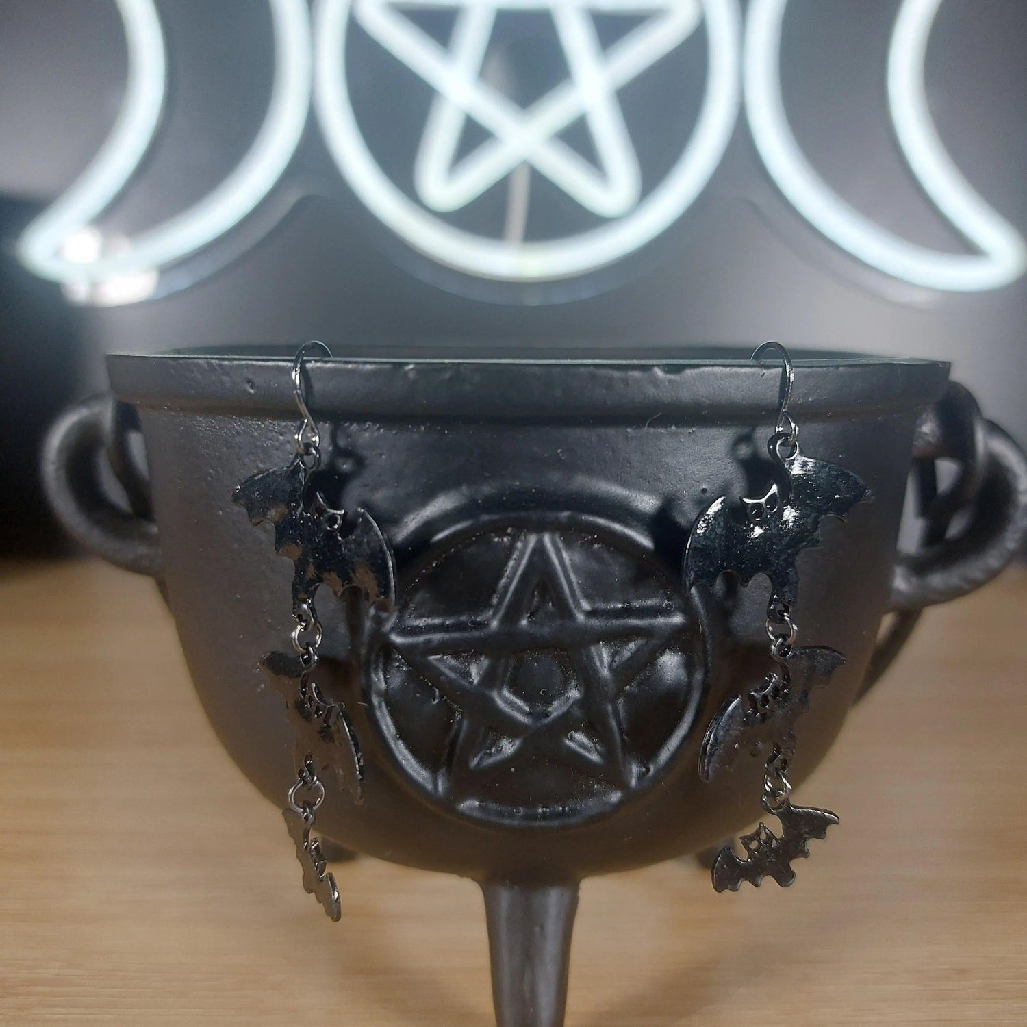 Black metal 3-tier bat dangle earrings hanging from a black cauldron with a pentagram.