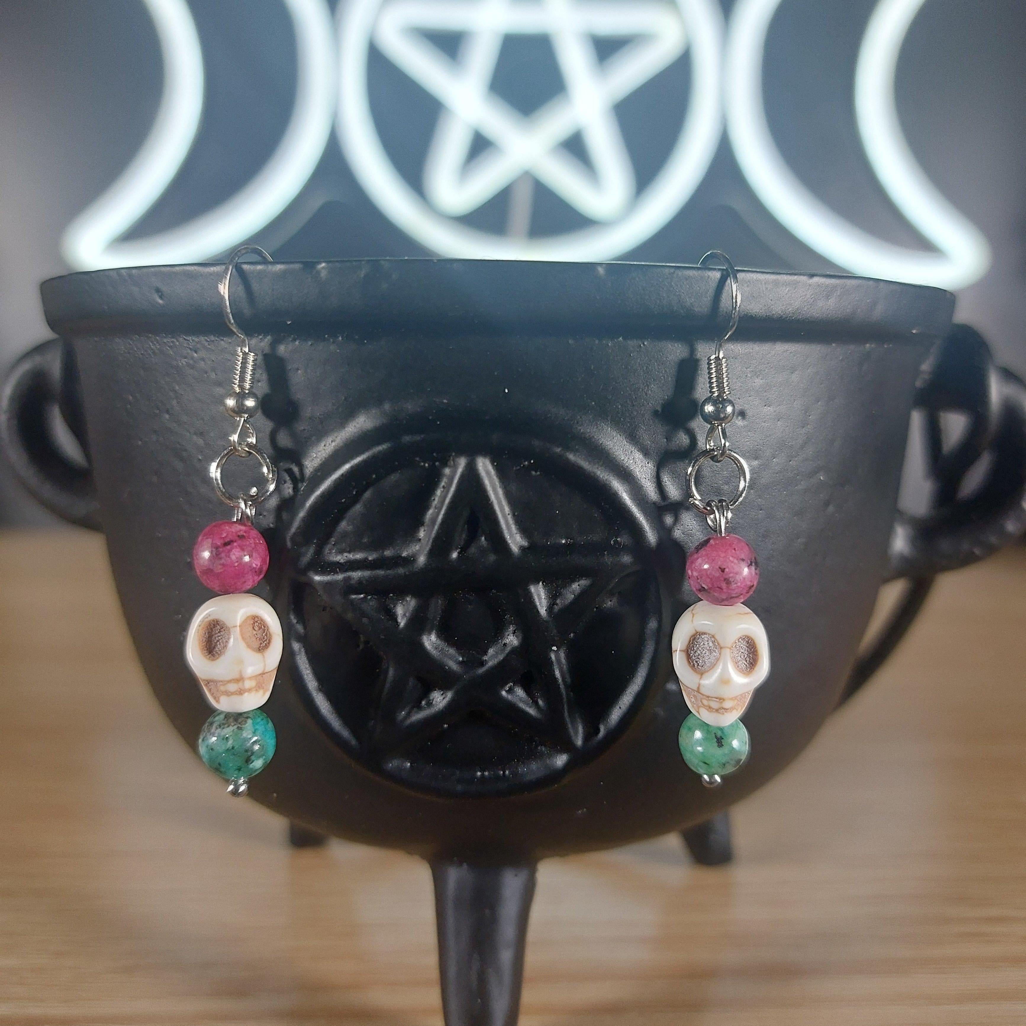 White howlite skull dangle earrings with pink and green glass beads on a black cauldron.