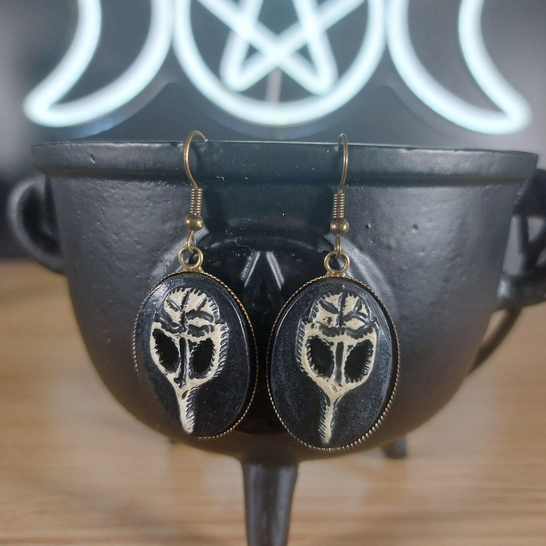 Antique bronze raven skull dangle earrings with black oval settings, hanging from a black cauldron.