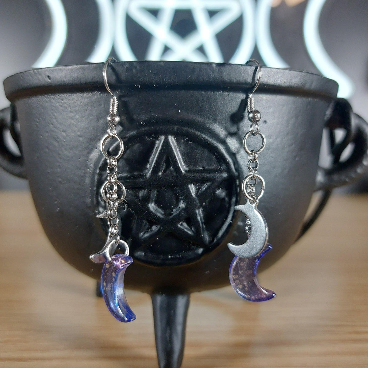 Purple glass moon dangle earrings with silver crescent moons, hanging from a black cauldron.