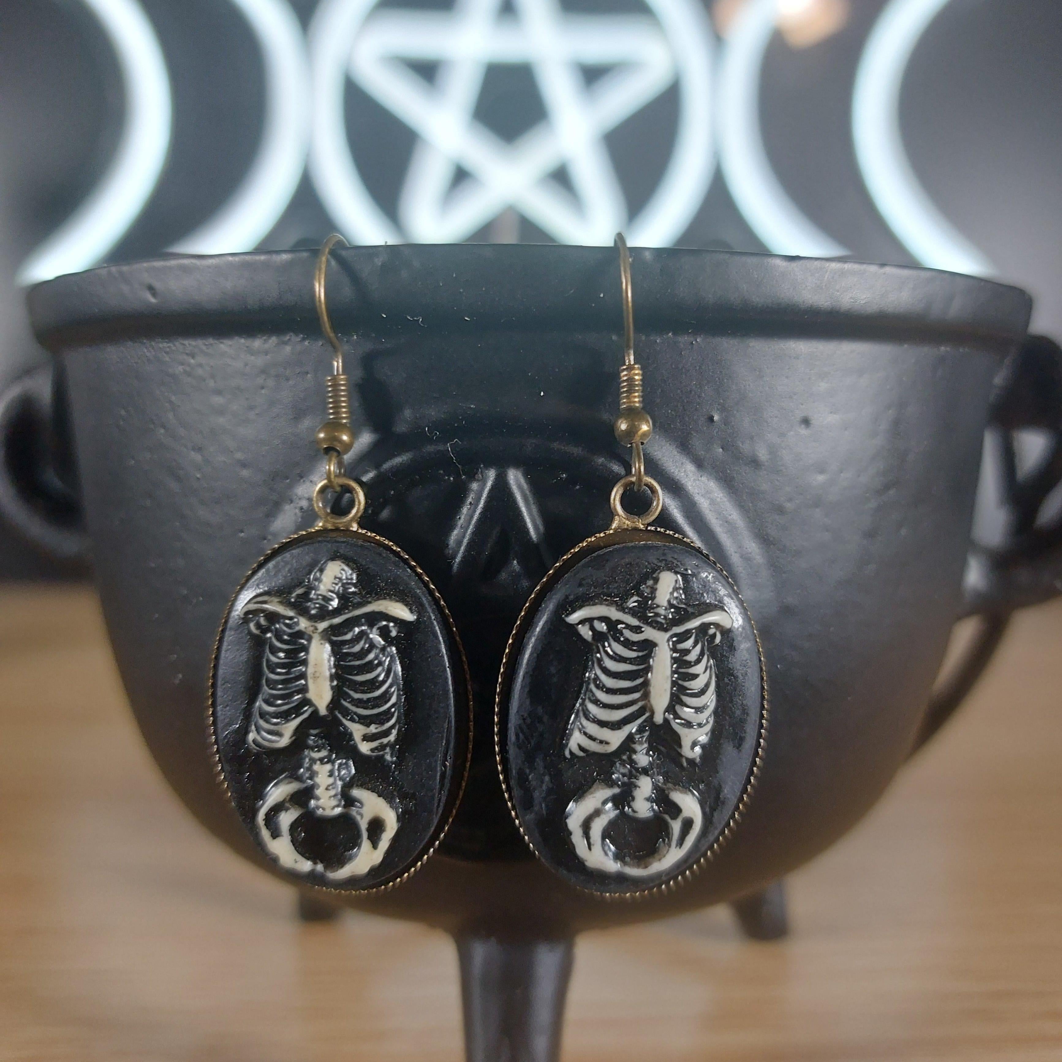 Antique bronze skeleton dangle earrings with black oval resin cabochons, displayed in a black cauldron.