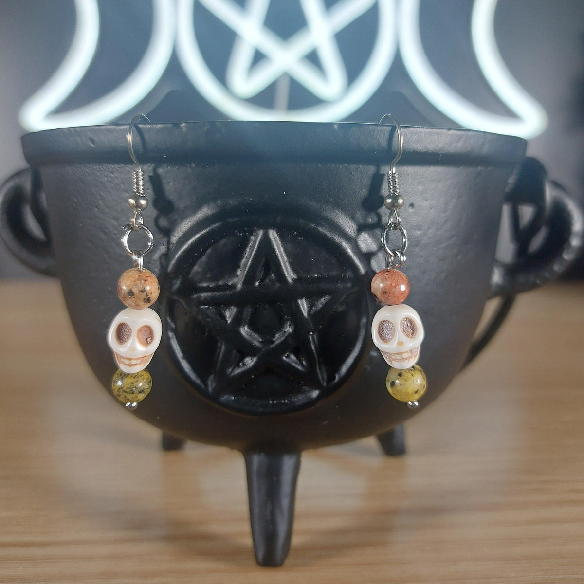 White howlite skull dangle earrings with yellow/orange glass beads, hanging from a black cauldron.