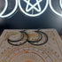 Black hollow metal crescent moon dangle earrings on an ornate book with a glowing triple moon symbol in the background.