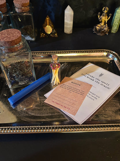 A ritual spell kit displayed on a silver tray, featuring a blue wand, a small glass jar of dried herbs, a matchstick holder with a miniature figure, and printed cards including one titled &