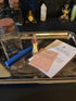 A ritual spell kit displayed on a silver tray, featuring a blue wand, a small glass jar of dried herbs, a matchstick holder with a miniature figure, and printed cards including one titled &