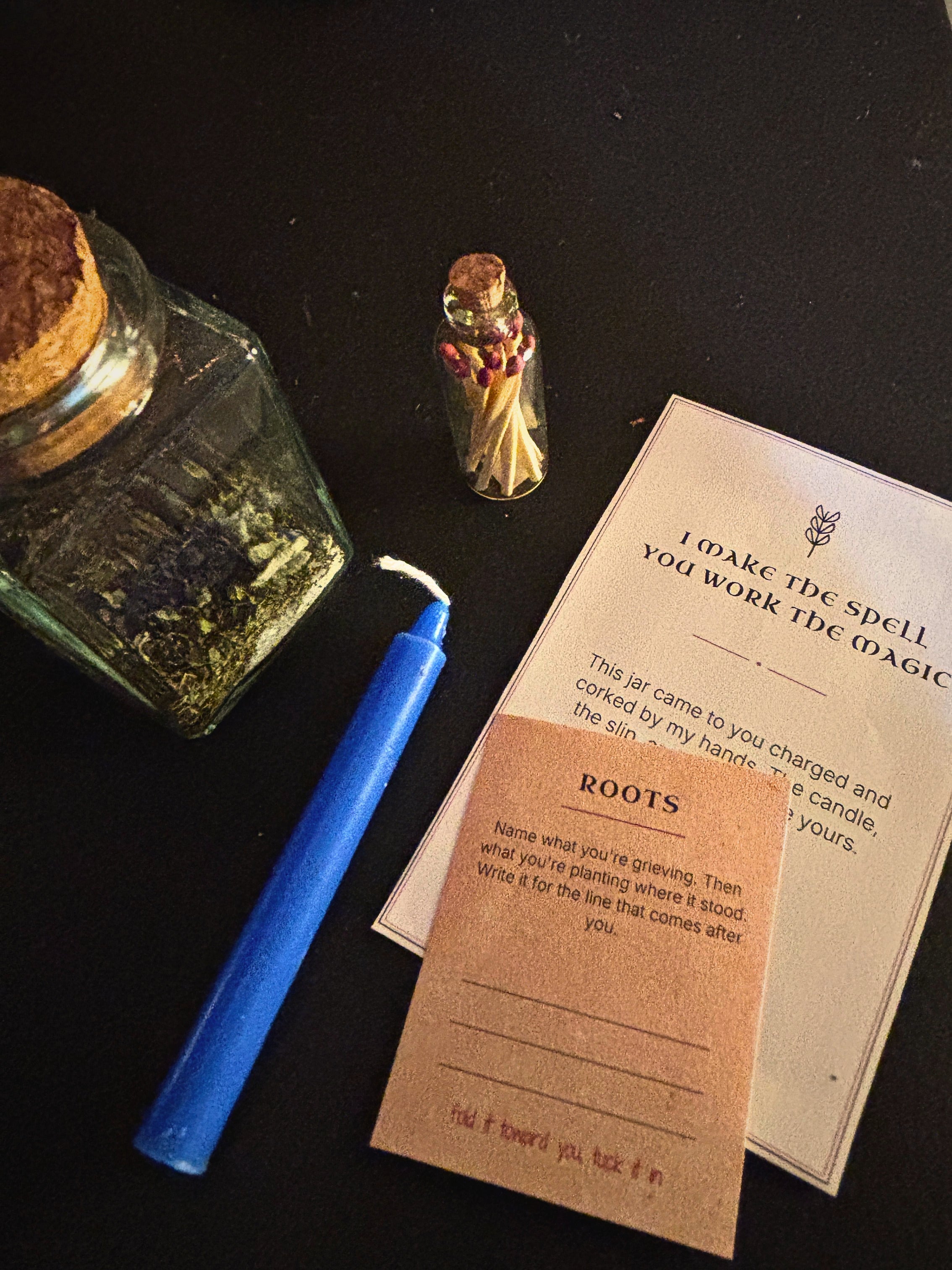 A flat-lay of a Wiccan altar kit featuring a blue taper candle, a small glass bottle of matches, a hexagram jar, and printed cards on a black background.